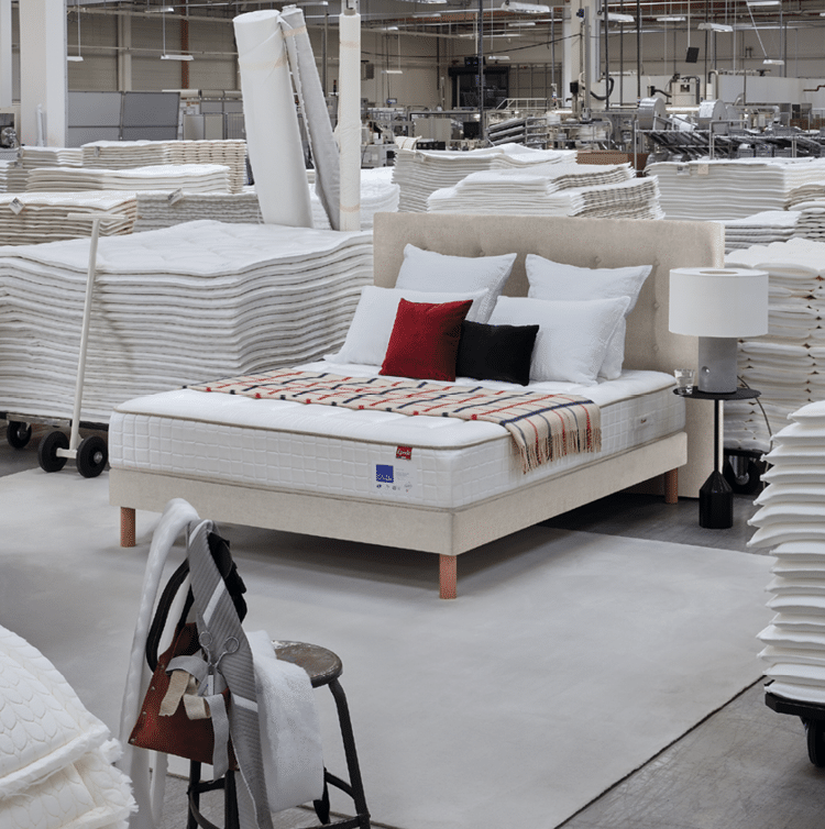 Un lit avec une t te de lit beige, des oreillers blancs et deux coussins color s est install dans un grand entrep t rempli de piles de tissus blancs et de machines industrielles.  Description g n r e par IA